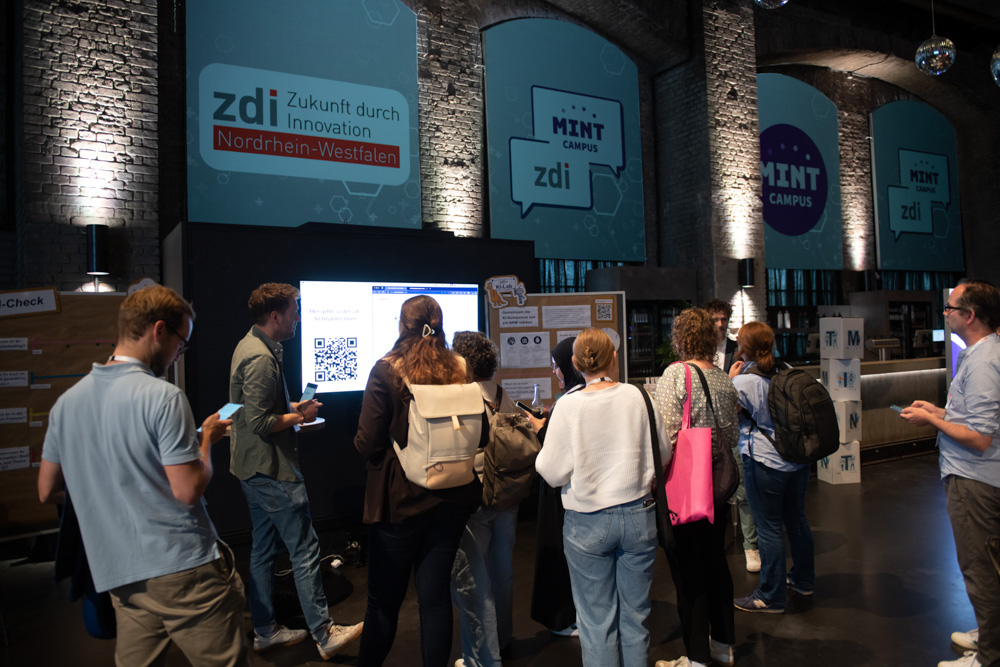 Eine Gruppe von Menschen steht bei einer Indoor-Veranstaltung vor einem Informationsdisplay und schwarzen Brettern mit Transparenten mit der Aufschrift "zdi" und "MINT CAMPUS".