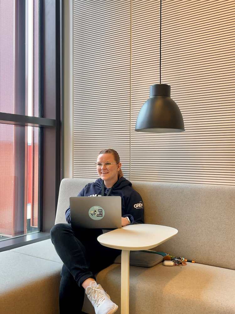 Eine Person sitzt auf einem beigen Sofa mit einem Laptop auf dem Schoß und lächelt. In der Nähe stehen ein kleiner weißer Tisch, eine schwarze Lampe und ein großes Fenster.