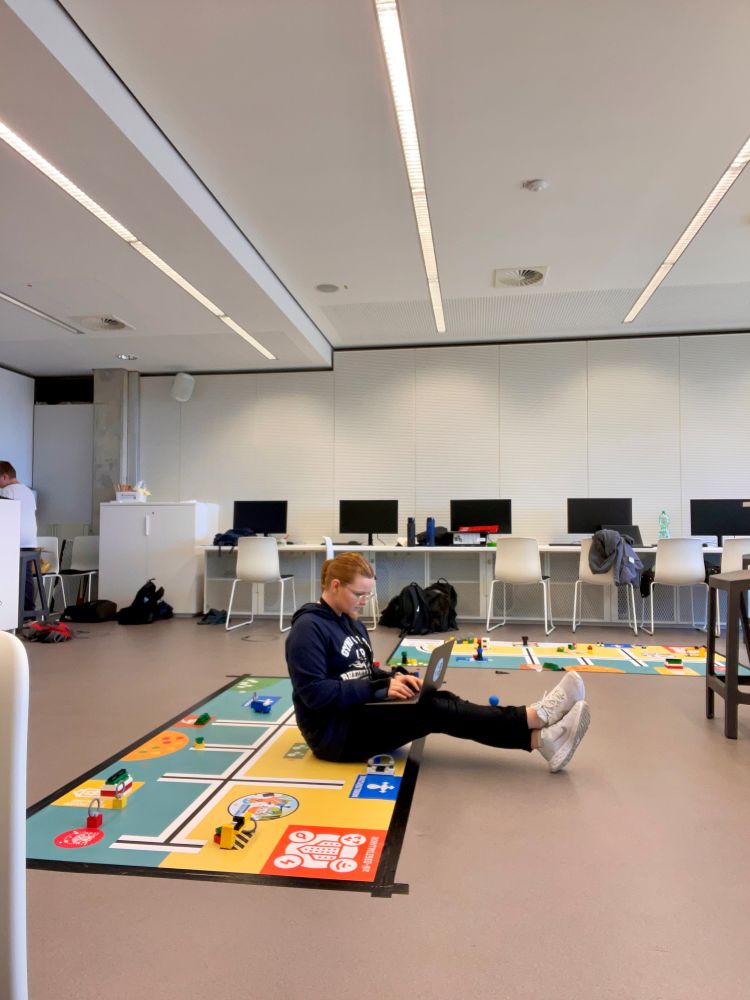 Eine Person sitzt mit einem Laptop in einem Klassenzimmer auf dem Boden. Im Hintergrund sind bunte Matten, Computer und Stühle zu sehen.