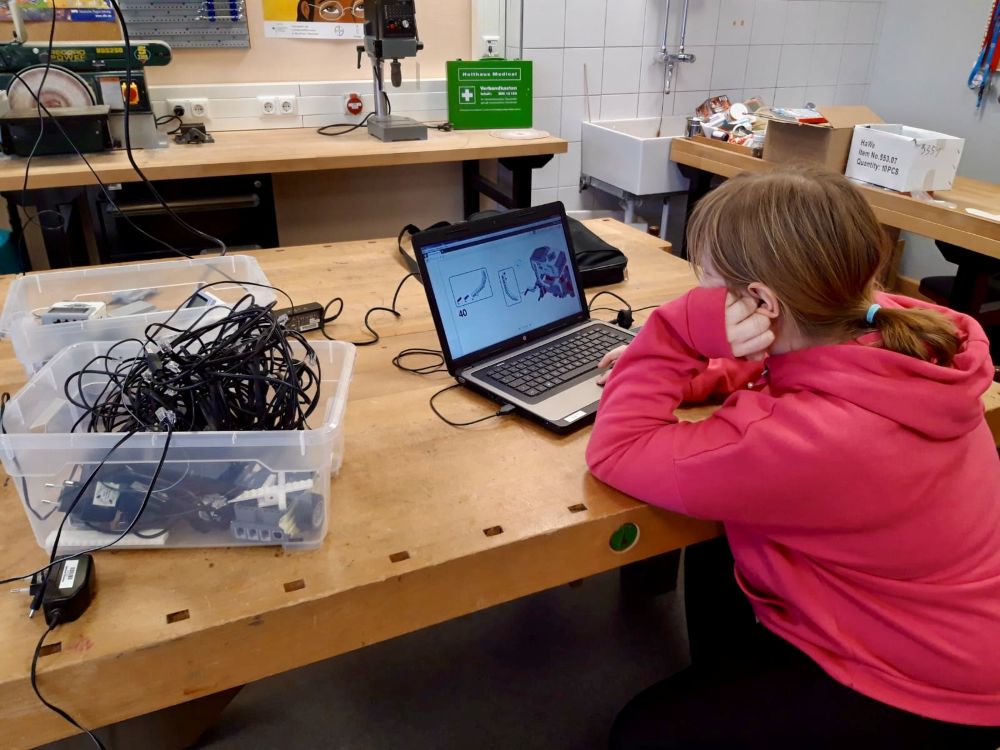 Mädchen im pinken Hoodie arbeitet an einem Laptop in einer Werkstatt mit Kabeln und Elektronikbauteilen