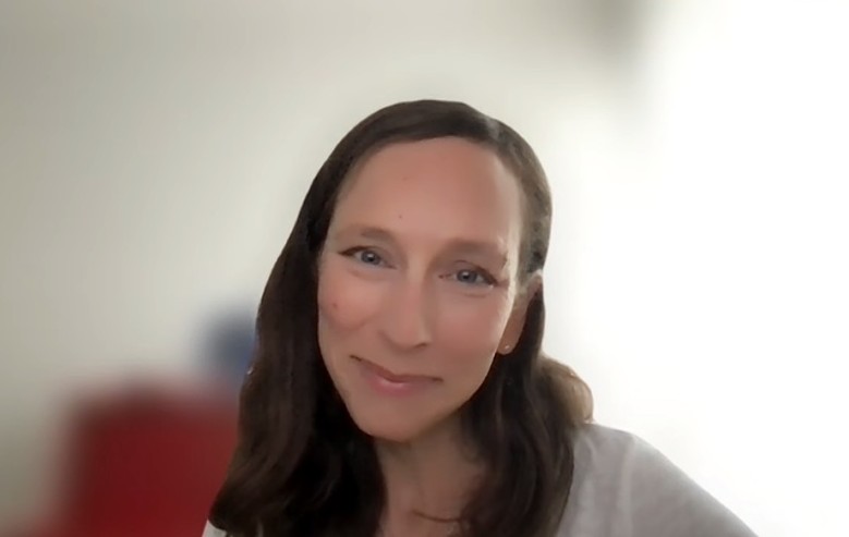 Die Frau hat langes braunes Haar und lächelt in die Kamera. Sie befindet sich in einem gut beleuchteten Raum mit einem unscharfen Hintergrund.