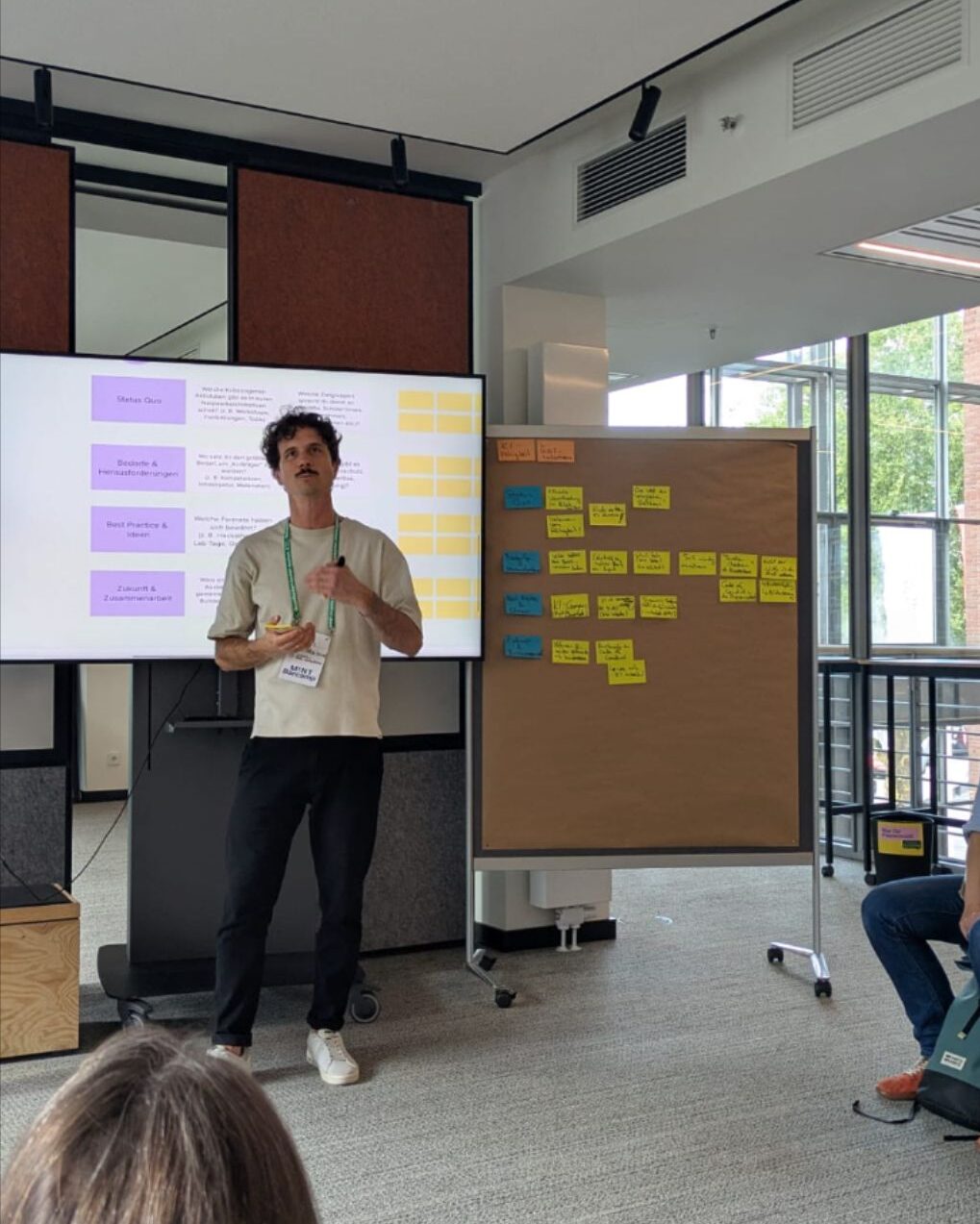Ein Mann mit Schnurrbart steht vor einem Bildschirm und einer Pinnwand mit Post-Its und gibt einen Workshop.