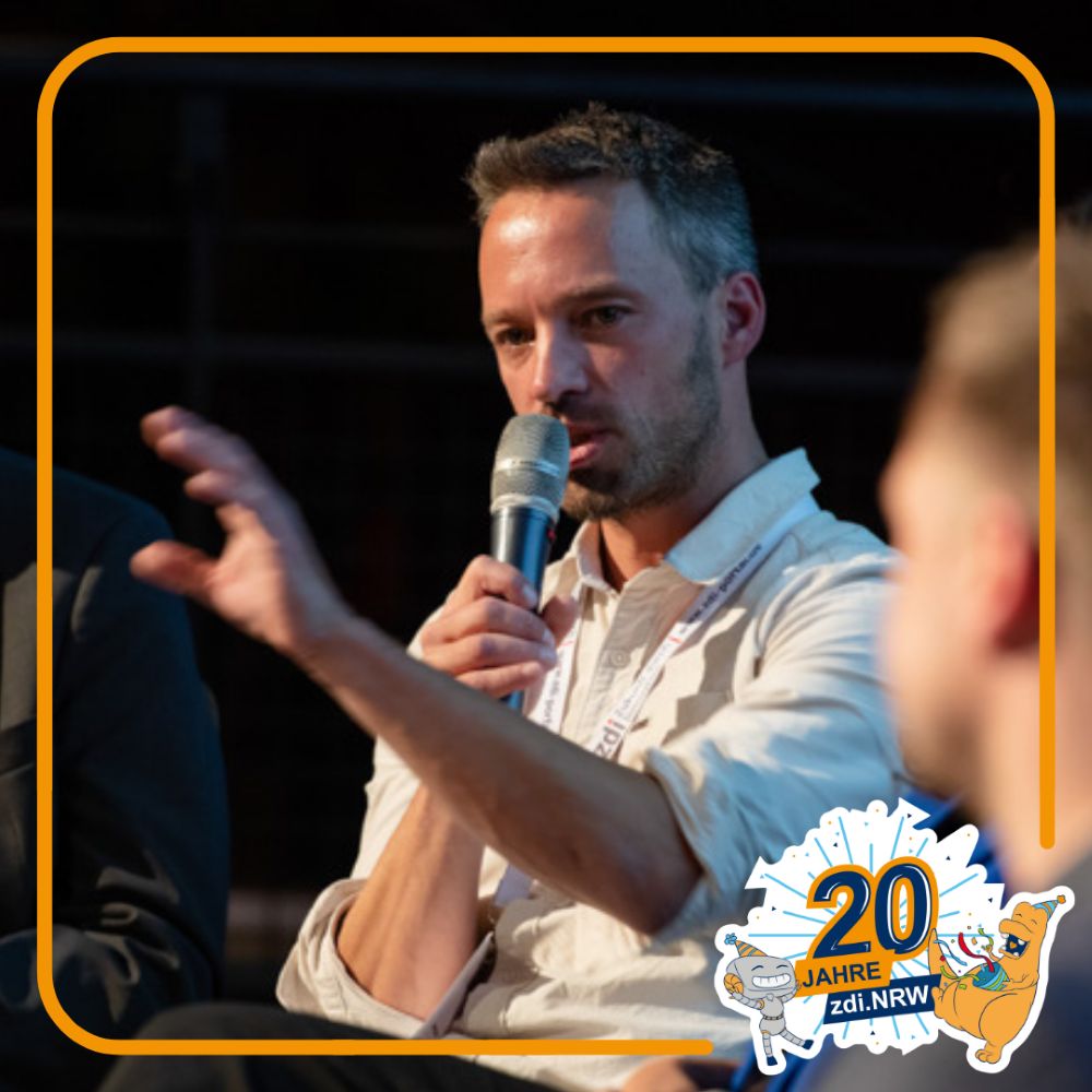 Ein Mann in einem hellen Hemd spricht während einer Podiumsdiskussion in ein Mikrofon. Rechts unten ist das Logo "20 Jahre zdi.NRW" zu sehen.