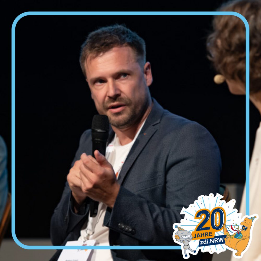 Ein Mann im Anzug spricht bei einer Podiumsdiskussion in ein Mikrofon. In der rechten unteren Ecke ist eine Grafik "20 Jahre zdi.NRW" zu sehen.