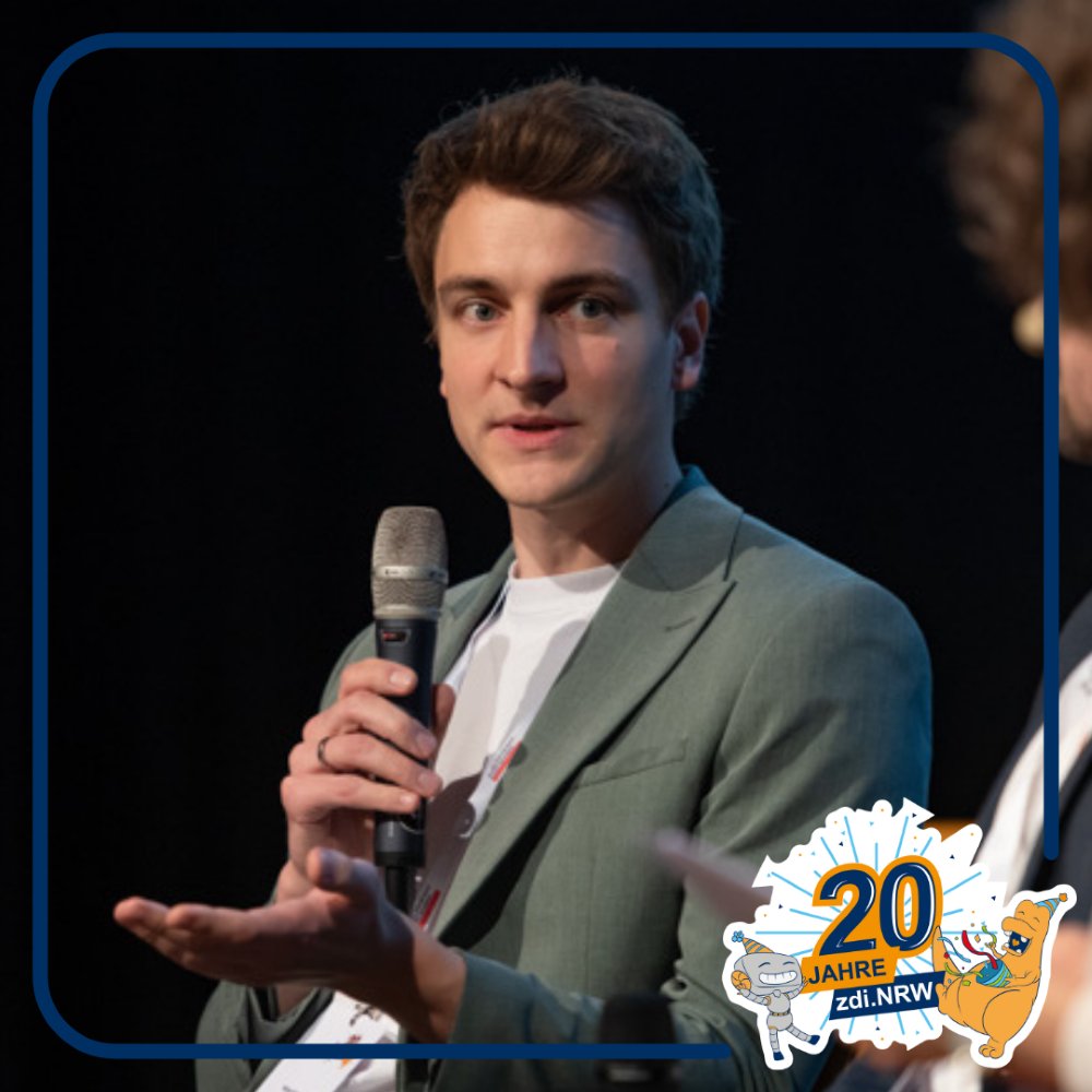 Eine Person in einem grünen Blazer hält ein Mikrofon und gestikuliert, während sie auf der Bühne spricht. Rechts unten erscheint das Logo "20 Jahre zdi.NRW".
