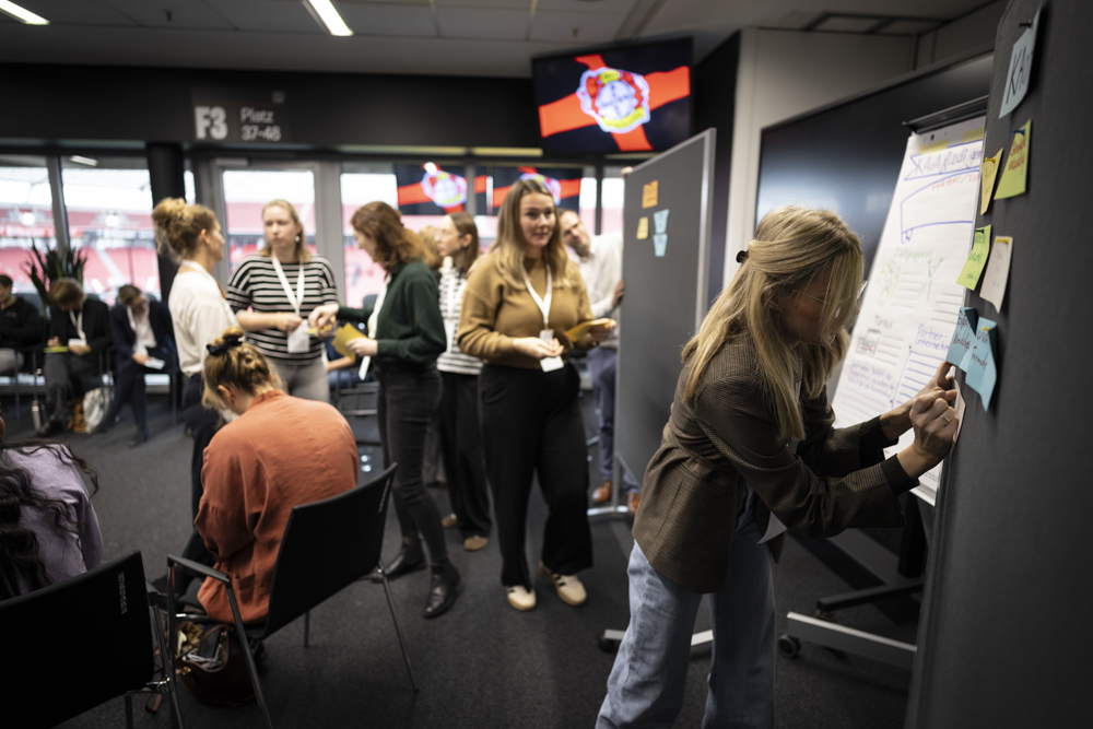 Menschen nehmen an einem Workshop teil. Einige schreiben auf Flipcharts und Haftnotizen, während andere in einem modernen Raum beobachten oder diskutieren.