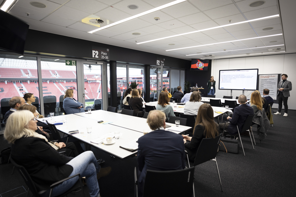 Menschen in Geschäftskleidung sitzen um Tische in einem Konferenzraum. Zwei Redner präsentieren vor einem großen Bildschirm. Durch die Fenster sind Stadionsitze zu sehen.