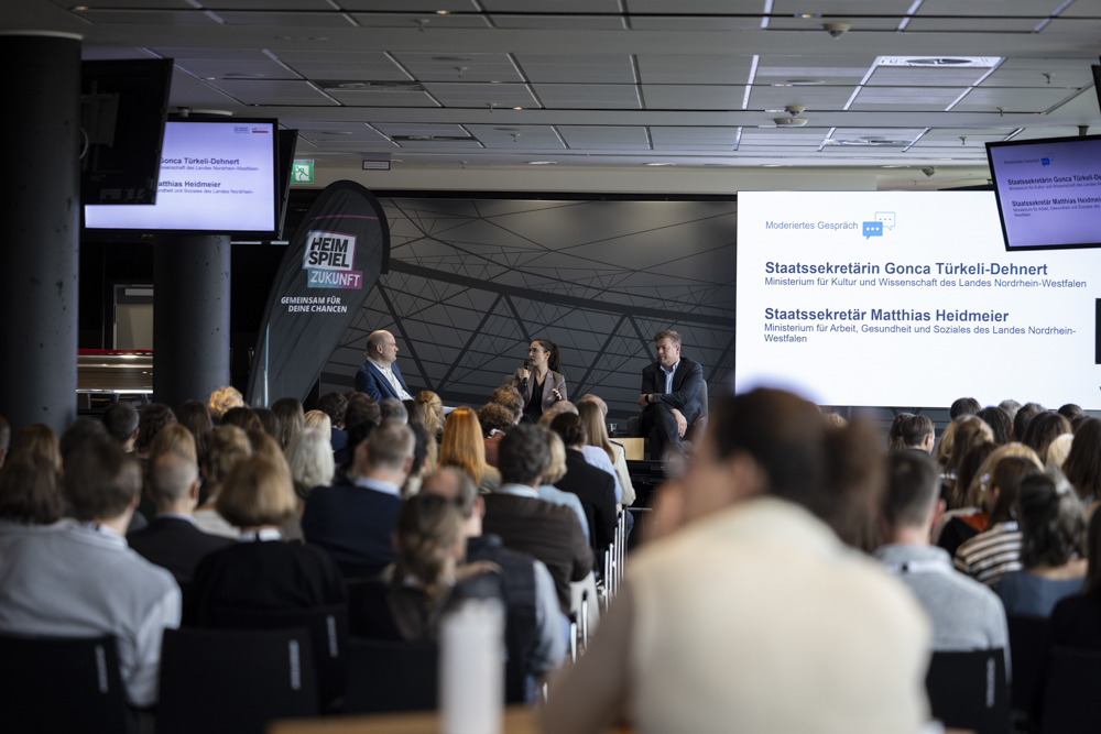 Eine Podiumsdiskussion findet auf der Bühne vor einem Publikum statt. Auf einem großen Bildschirm hinter ihnen werden Informationen über die Redner angezeigt.