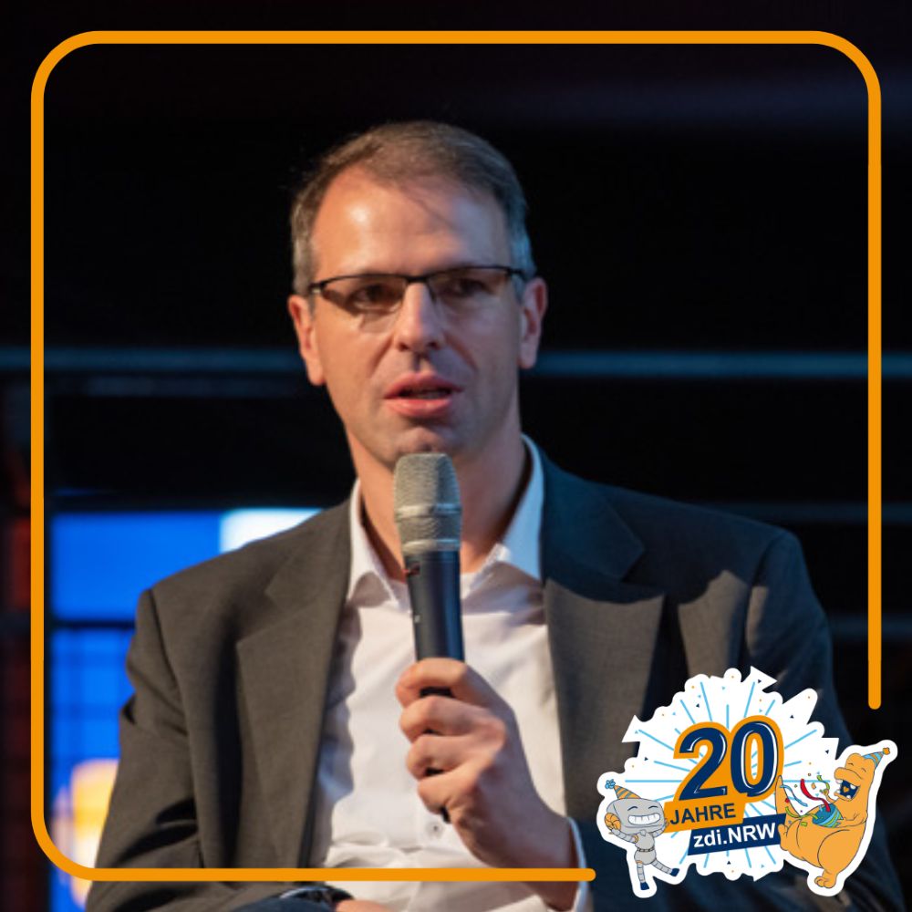 Ein Mann im Anzug und mit Brille spricht in ein Mikrofon. In der rechten unteren Ecke erscheint das Logo "20 Jahre zdi.NRW".