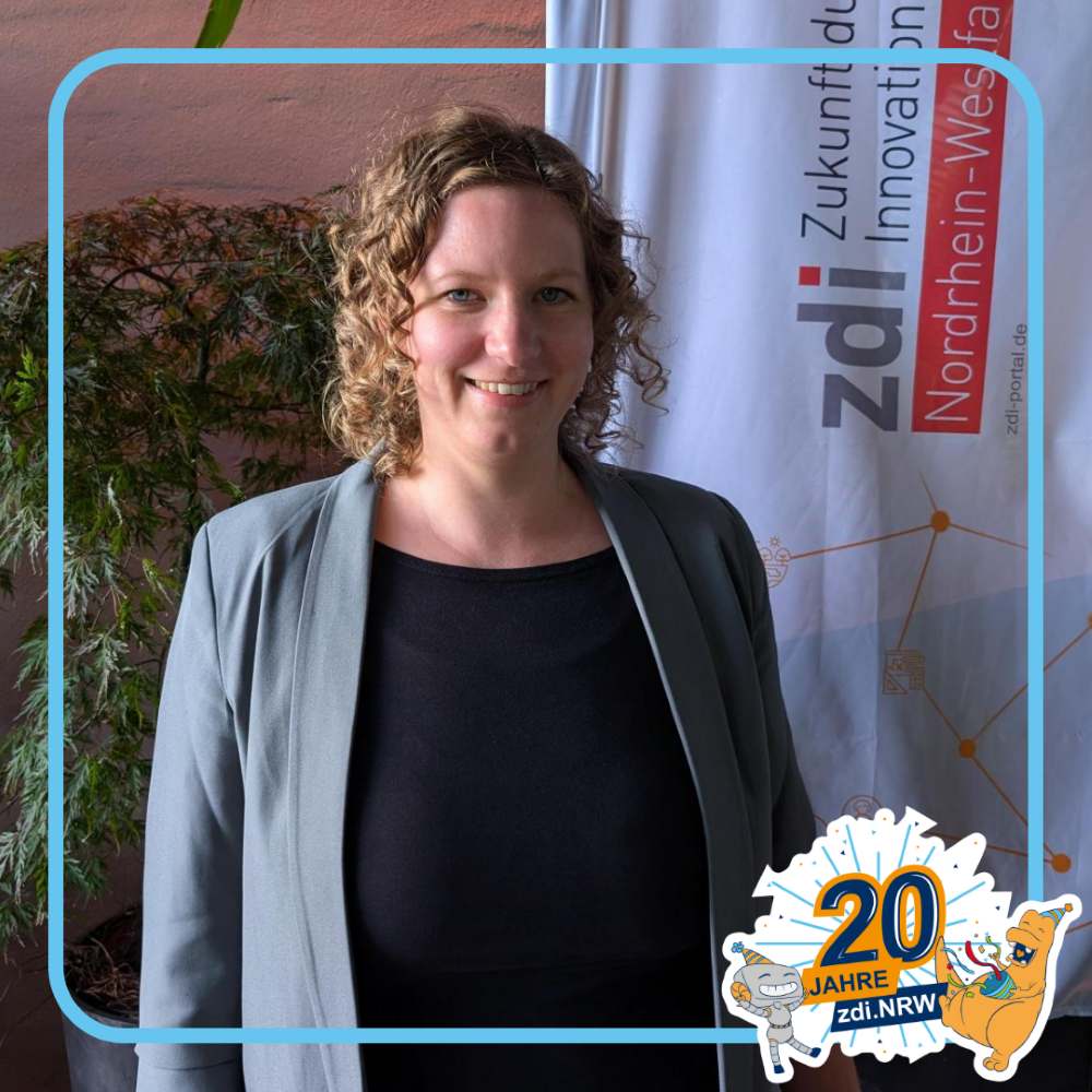 Eine Frau mit lockigem Haar steht in einem Haus und lächelt vor einem zdi.NRW-Banner. Rechts unten erscheint das Logo "20 Jahre zdi.NRW".