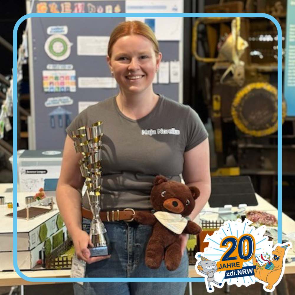 Eine junge Frau steht mit einer Trophäe und einem braunen Teddybär in der Hand vor einem Messestand zum 20-jährigen Bestehen von zdi.NRW.