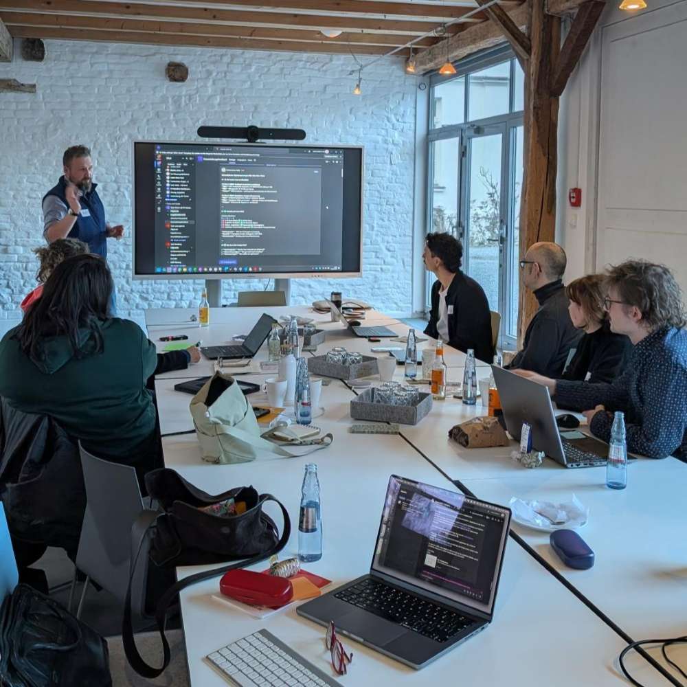 Workshop in einem Besprechungsraum: Eine Person präsentiert an einem großen Bildschirm, mehrere Teilnehmende sitzen am Tisch mit Laptops