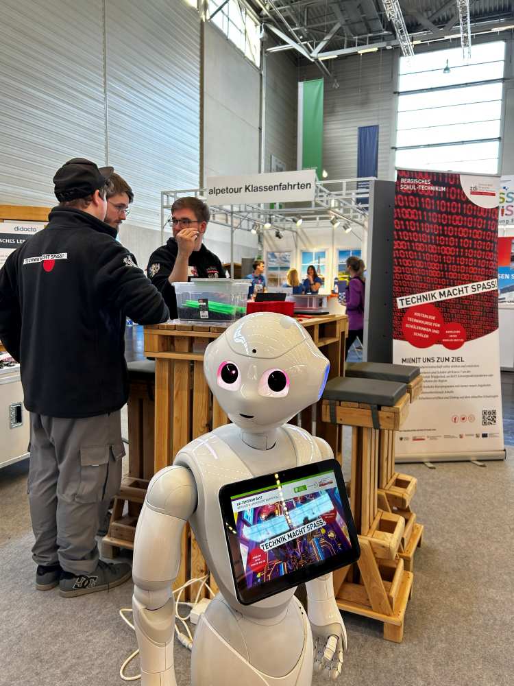Ein humanoider Roboter mit einem Touchscreen steht vor einem Stand. Zwei Personen unterhalten sich neben einem Holztresen auf einer Technologieausstellung.