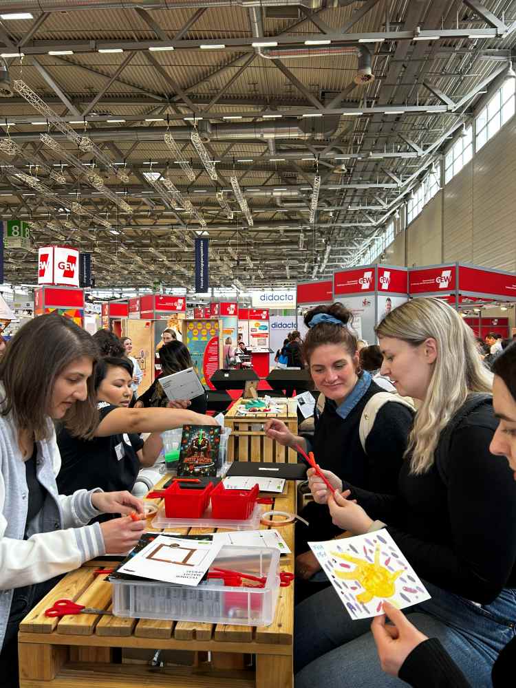 Frauen sitzen auf einem Kongress an einem Tisch, während sie an Kunsthandwerk arbeiten und sich Kunstzubehör ansehen. Hinter ihnen sind Stände und Auslagen zu sehen.