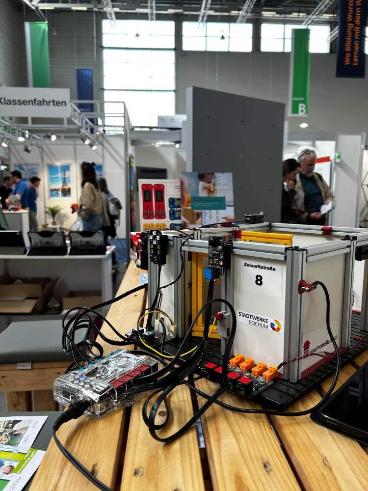 Ein Modell einer Energiestation mit Drähten und Sensoren steht auf einem Holztisch in einer Ausstellung. Im Hintergrund sind Menschen und Stände zu sehen.