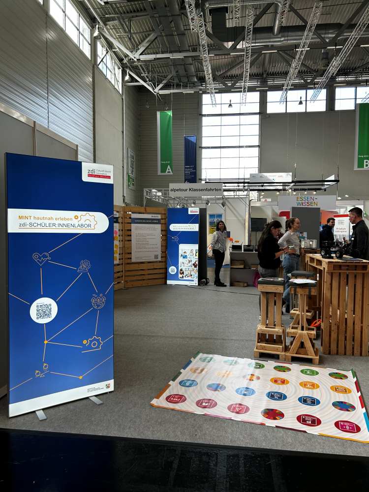 Ein Messestand mit Informationsbannern, einem Holztresen mit Menschen in der Nähe und einer bunten Matte mit verschiedenen Symbolen.