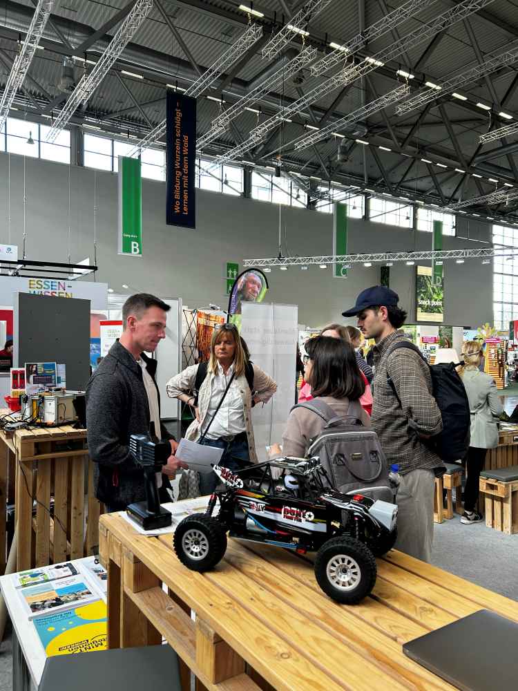 Eine Gruppe von Menschen steht um einen Stand auf einer Indoor-Ausstellung. Ein ferngesteuertes Auto steht auf einem Holztisch davor.