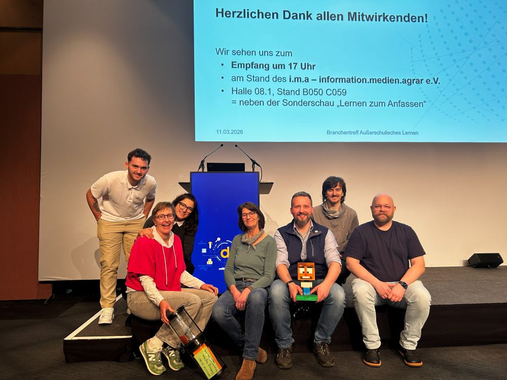 Sieben Personen stehen gemeinsam auf einer Bühne. Hinter ihnen zeigt ein Bildschirm Veranstaltungshinweise und Danksagungen in deutscher Sprache.