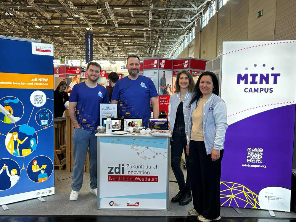 Vier Personen stehen an einem Stand von zdi.NRW und MINT-Campus auf einer Indoor-Veranstaltung. Um sie herum sind Banner und Materialien ausgelegt.