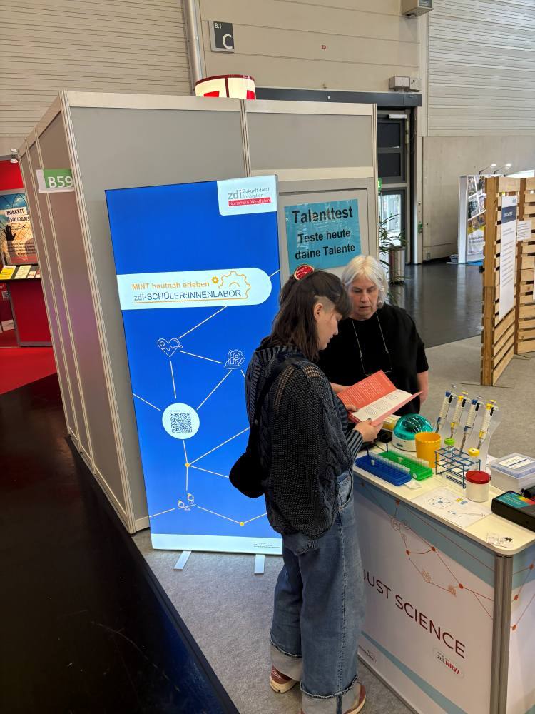 Zwei Frauen stehen an einem Stand mit Informationsmaterial, einem Reagenzglasständer und einem "Talenttest"-Schild auf dem Tisch.
