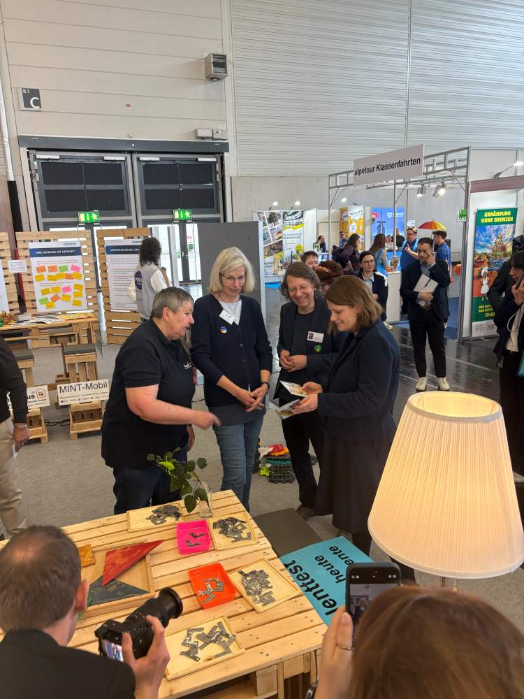 Eine Gruppe von Personen steht bei einer Indoor-Veranstaltung um einen Ausstellungstisch herum. Sie betrachten Materialien und interagieren, während andere im Hintergrund zu sehen sind.