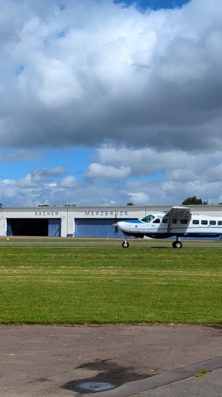 Auf der Wiese des Flugplatzes, vor einem Hangar mit der Aufschrift „Aachen Merzbrück“, steht unter einem bewölkten Himmel ein kleines Flugzeug.