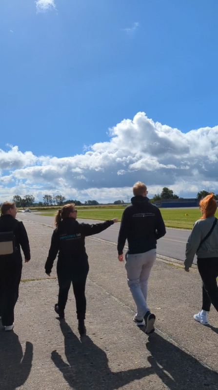 Vier Menschen gehen unter einem strahlend blauen Himmel mit vereinzelten Wolken auf einem gepflasterten Weg am Flugplatz entlang. Eine der Menschen deutet in die Ferne.