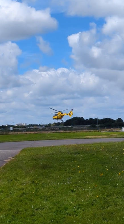 Ein gelber Hubschrauber fliegt tief über eine Wiese in der Nähe des Flugplatzes. Der Himmel ist teilweise bewölkt. Im Vordergrund verläuft ein gepflasterter Weg.
