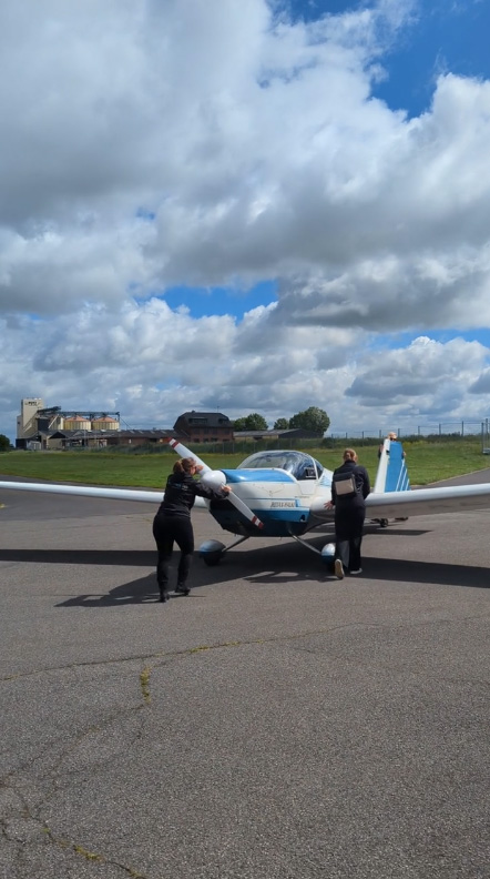 Zwei Personen schieben unter einem teilweise bewölkten Himmel ein kleines blau-weißes Flugzeug über das Rollfeld eines Flugplatzes.