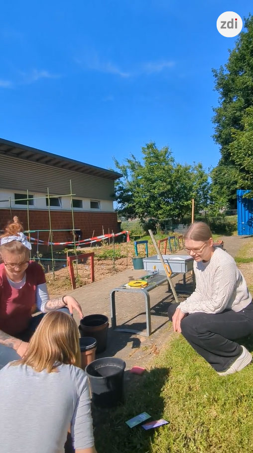 Drei Personen gärtnern im Freien mit Pflanzen, Töpfen und Gartengeräten. Im Hintergrund sind unter einem klaren blauen Himmel ein Gebäude und Bäume zu sehen.