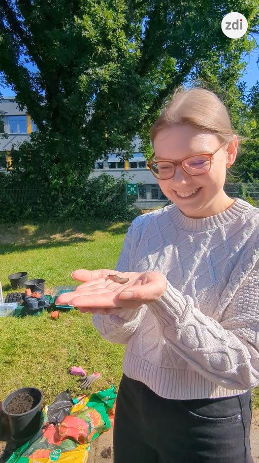 Eine Person mit Brille und leichtem Pullover lächelt im Freien und hält dabei eine kleine Schnecke in der Hand. Im Hintergrund sind Gartenutensilien zu sehen.
