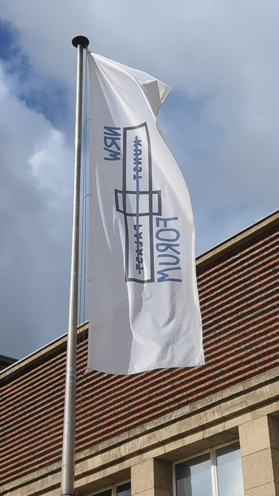 Weiße Fahne mit dem Logo von IWM und dem Schriftzug „Kunst“ vor einem Gebäude im Wind