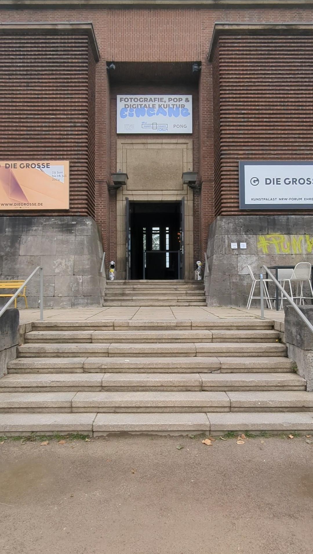 Eingang eines Backsteingebäudes mit Treppe und Ausstellungsschildern zu „Die Große“ und „Fotografie, Pop & Digitale Kultur“