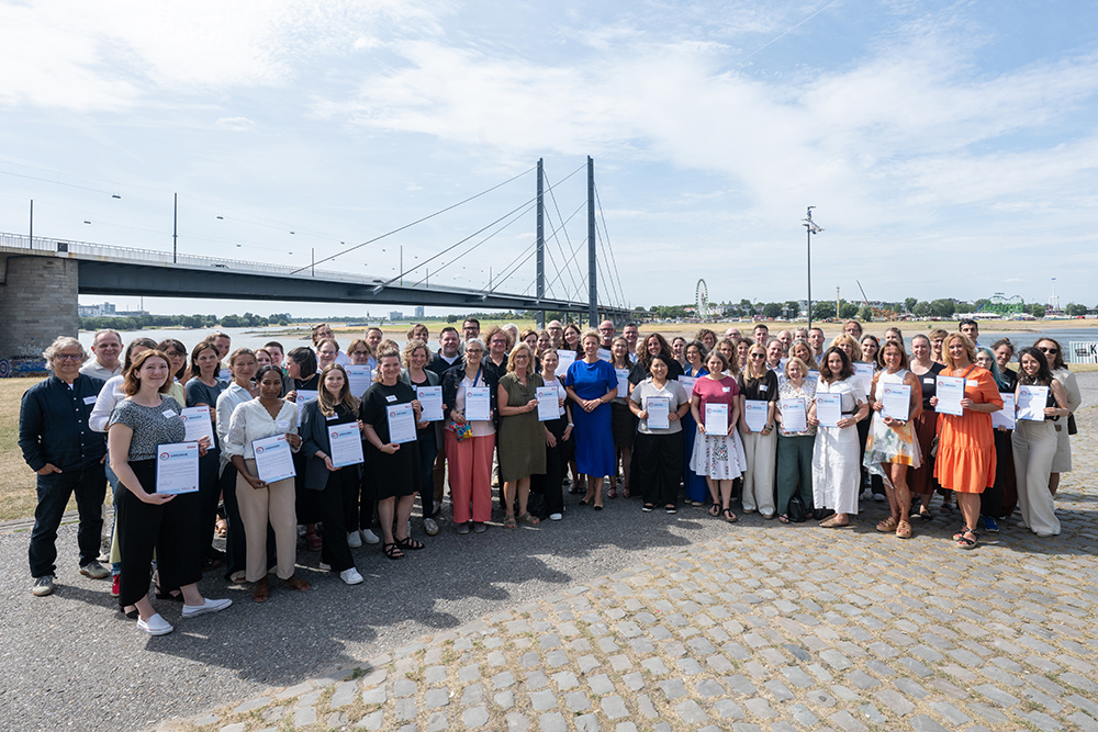 Gruppenfoto von rund 50 Personen mit Urkunden vor einer Brücke am Flussufer