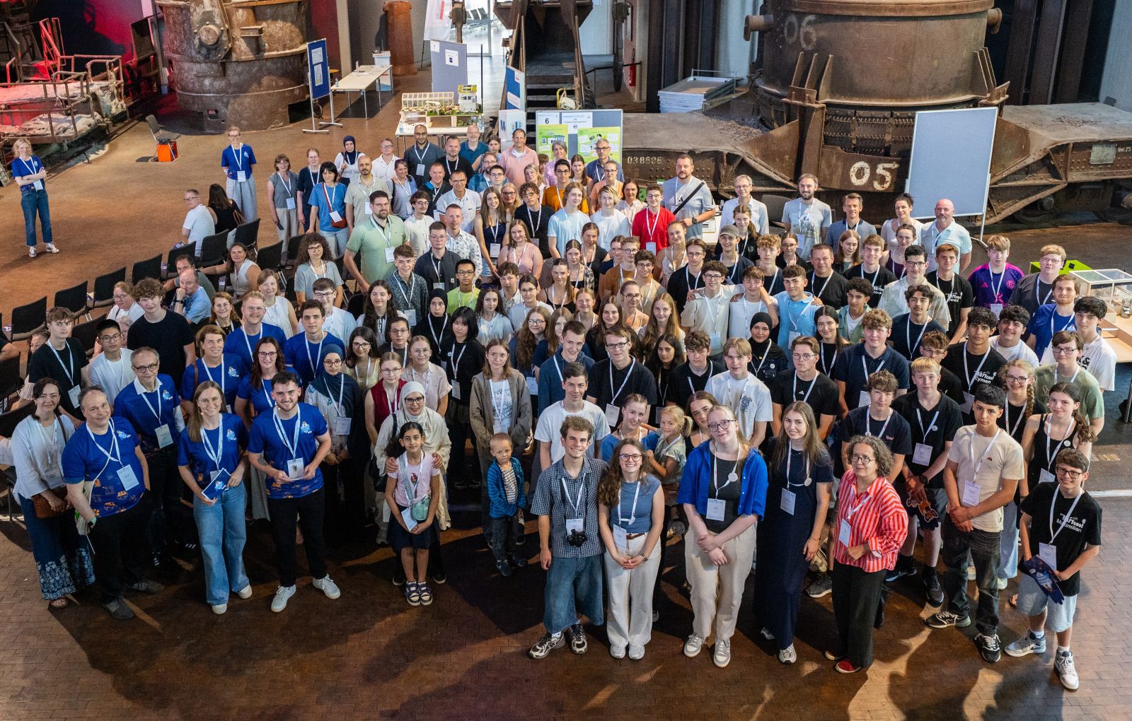 Gruppenfoto von vielen Teilnehmenden mit Namensschildern in einer Industriehalle mit großen Maschinen im Hintergrund
