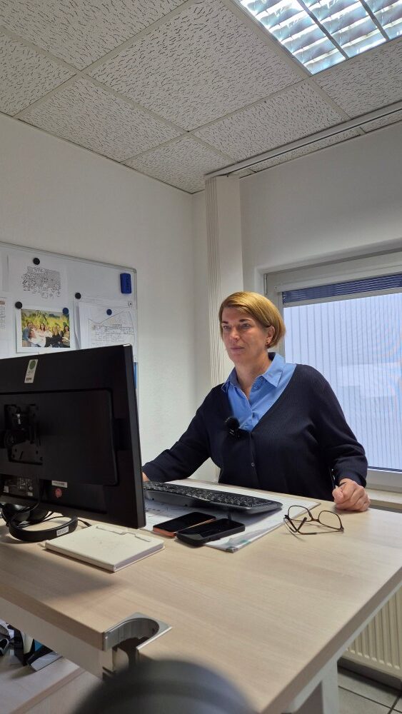 Eine Frau sitzt an einem Schreibtisch mit einem Computer in einem modernen Büro. Im Hintergrund sind ein Whiteboard und ein Fenster zu sehen.