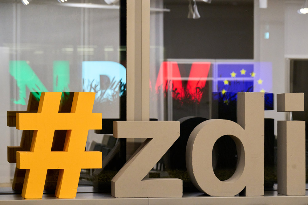 Ein Schild mit der Aufschrift „zdi“ vor einem Fenster. Durch das Fenster sieht man die Buchstaben NRW in Grün, Weiß und Rot und eine Europafahne leuchten.