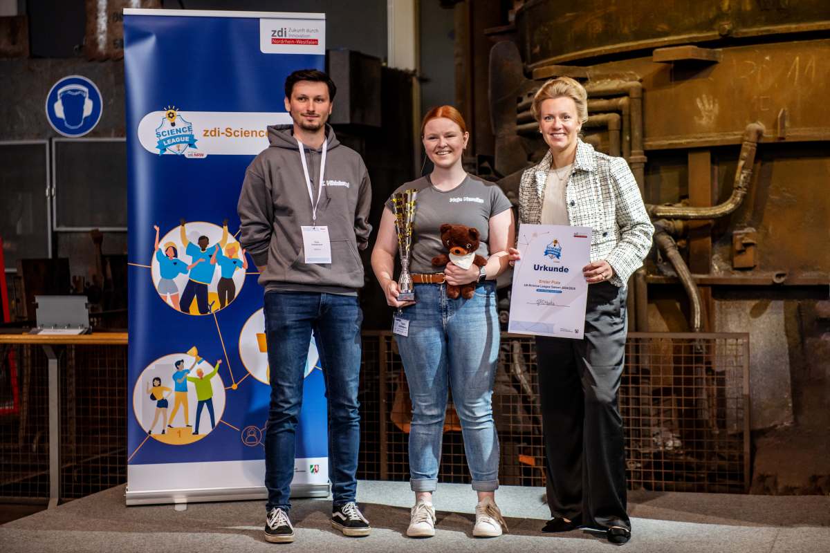 Drei Personen stehen auf einer Bühne. Die Person in der Mitte hält eine Trophäe und einen Teddybären in den Händen. Die Person rechts hält eine Urkunde. Im Hintergrund ist ein zdi-Science-Banner zu sehen.