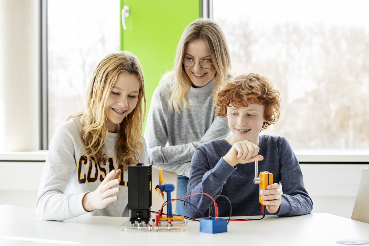 Das Foto zeigt ein Mädchen und einen Jungen, die ein Experiment mit einem Windrad durchführen. Eine Frau schaut ihnen über die Schulter und lächelt ermutigend.