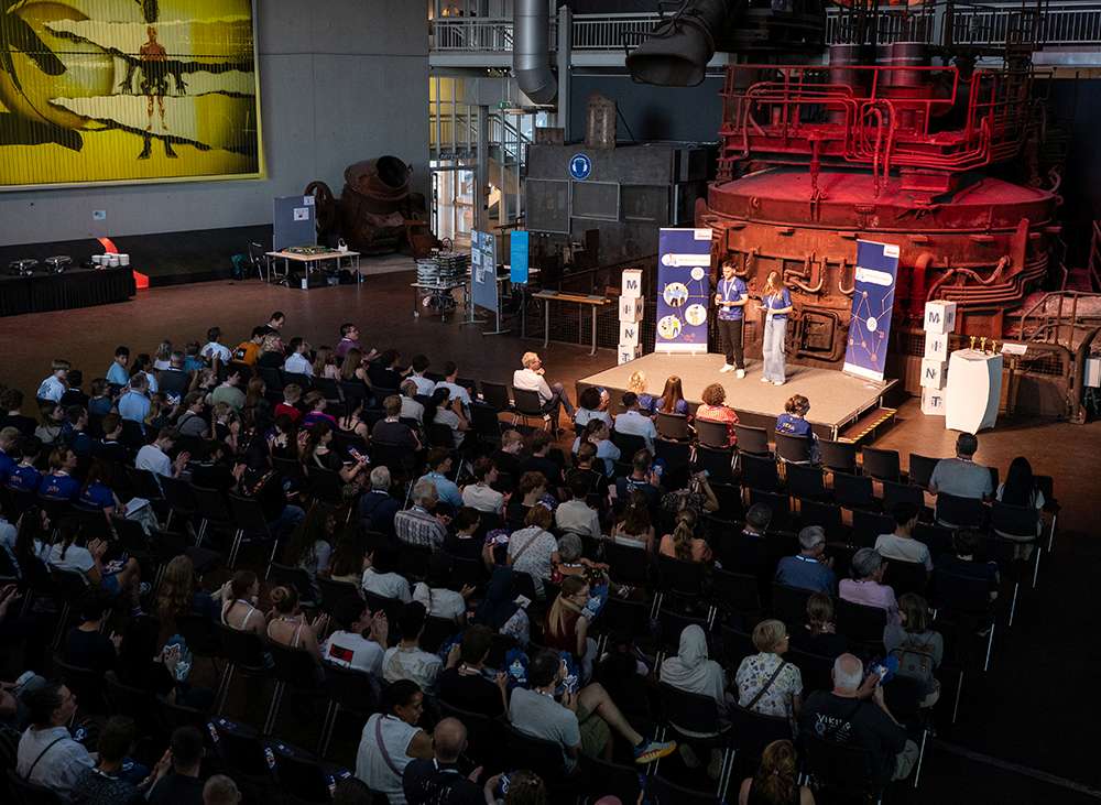 Vortrag vor großem Publikum in einer Industriehalle mit Bühne und roter Maschinenanlage