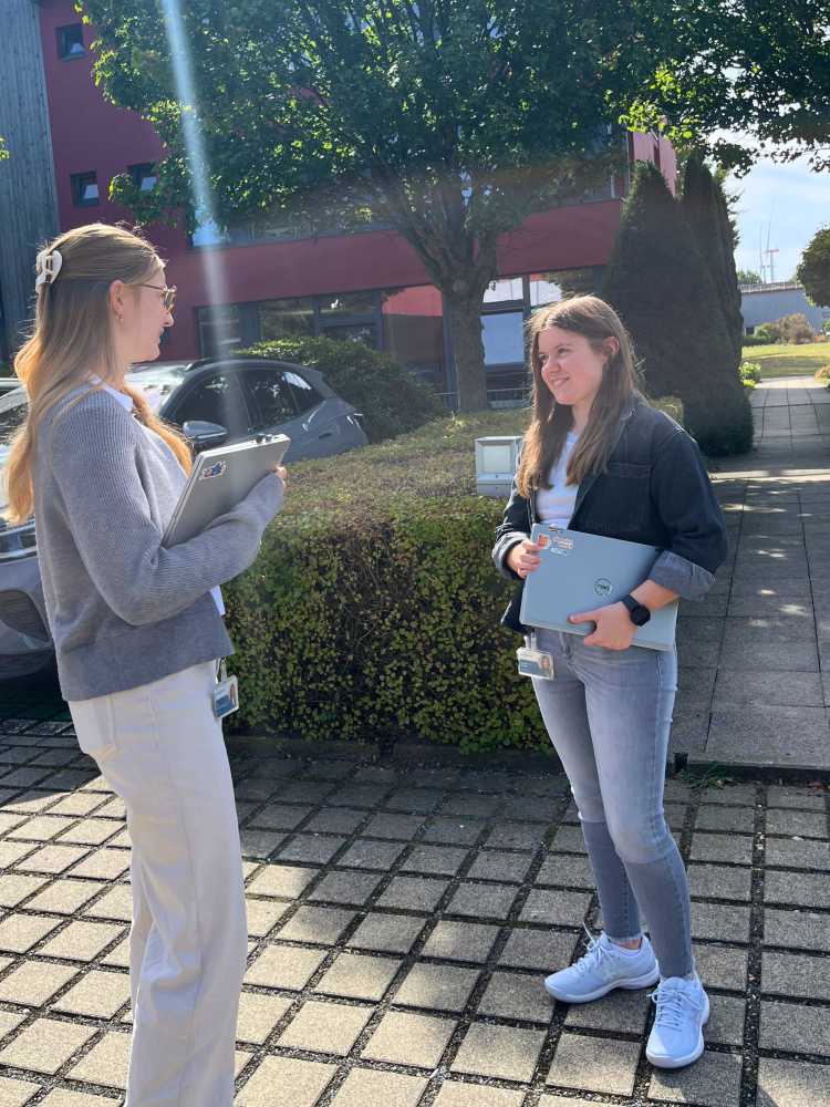 Vanessa Kriebel steht vor einem Gebäude und spricht mit einer Kollegin. Unter dem Arm hält sie einen Laptop.